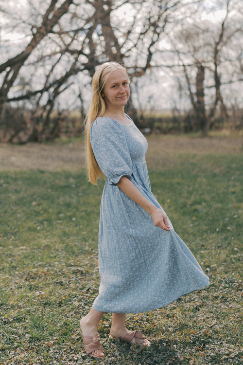 Woman in a blue midi dress standing in a field with bare trees in the background